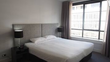 A hotel room with a large bed, two bedside tables with lamps, a window with curtains, and a view of buildings outside.