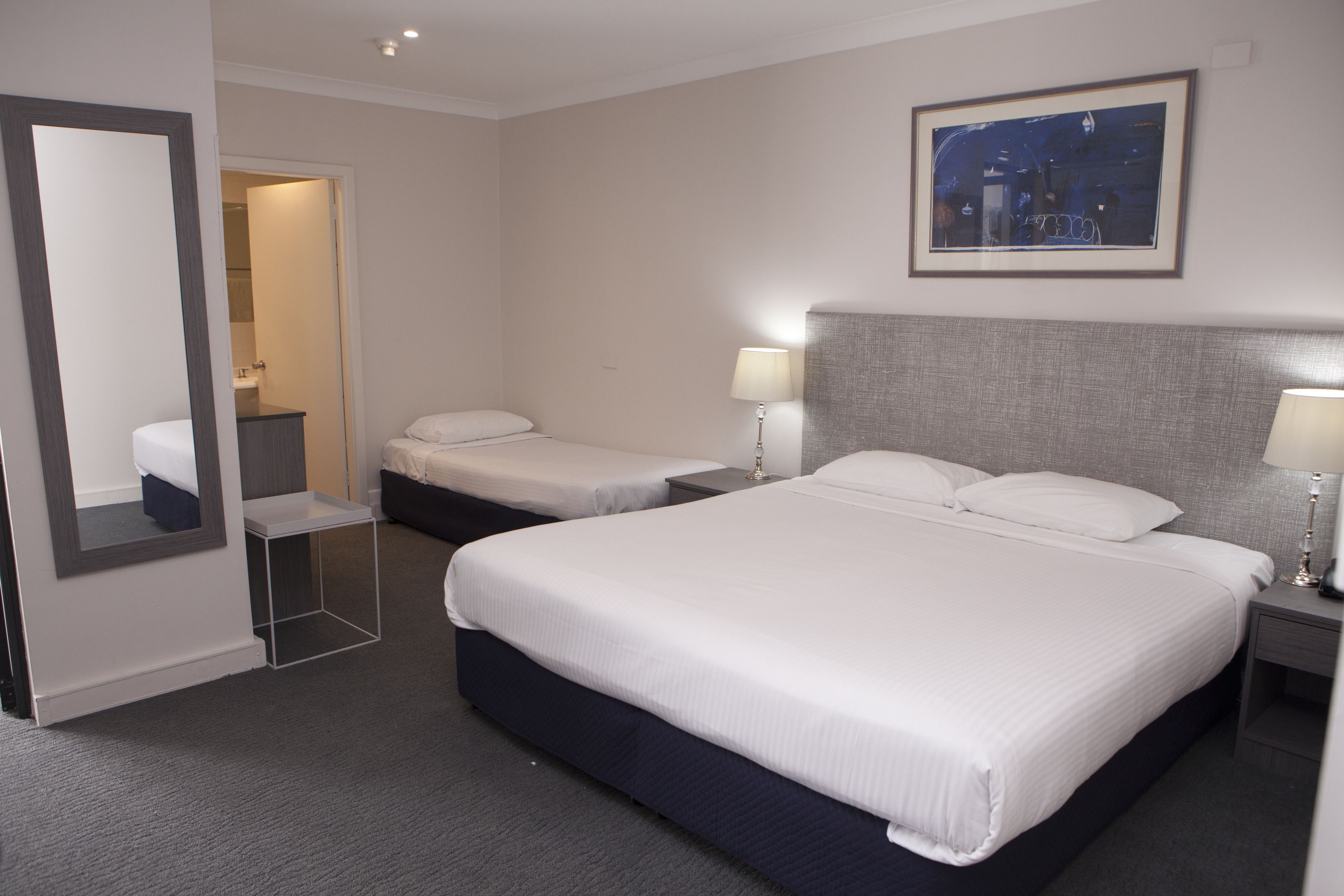 Habitación de hotel con dos camas, un espejo, un cuadro enmarcado en la pared y una mesita de noche con una lámpara.