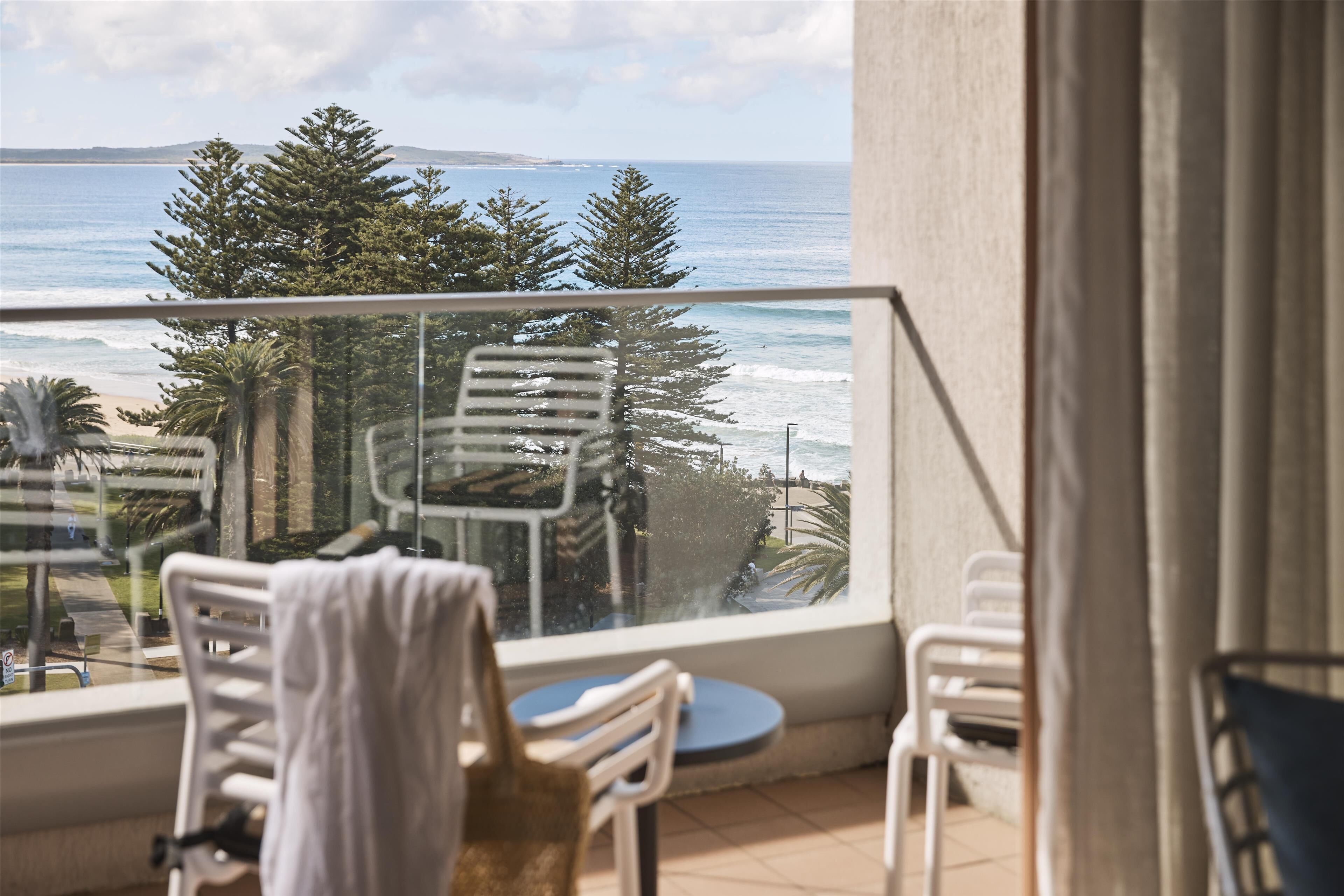 A balcony with a view of the ocean and beach, featuring white chairs and a small round table.