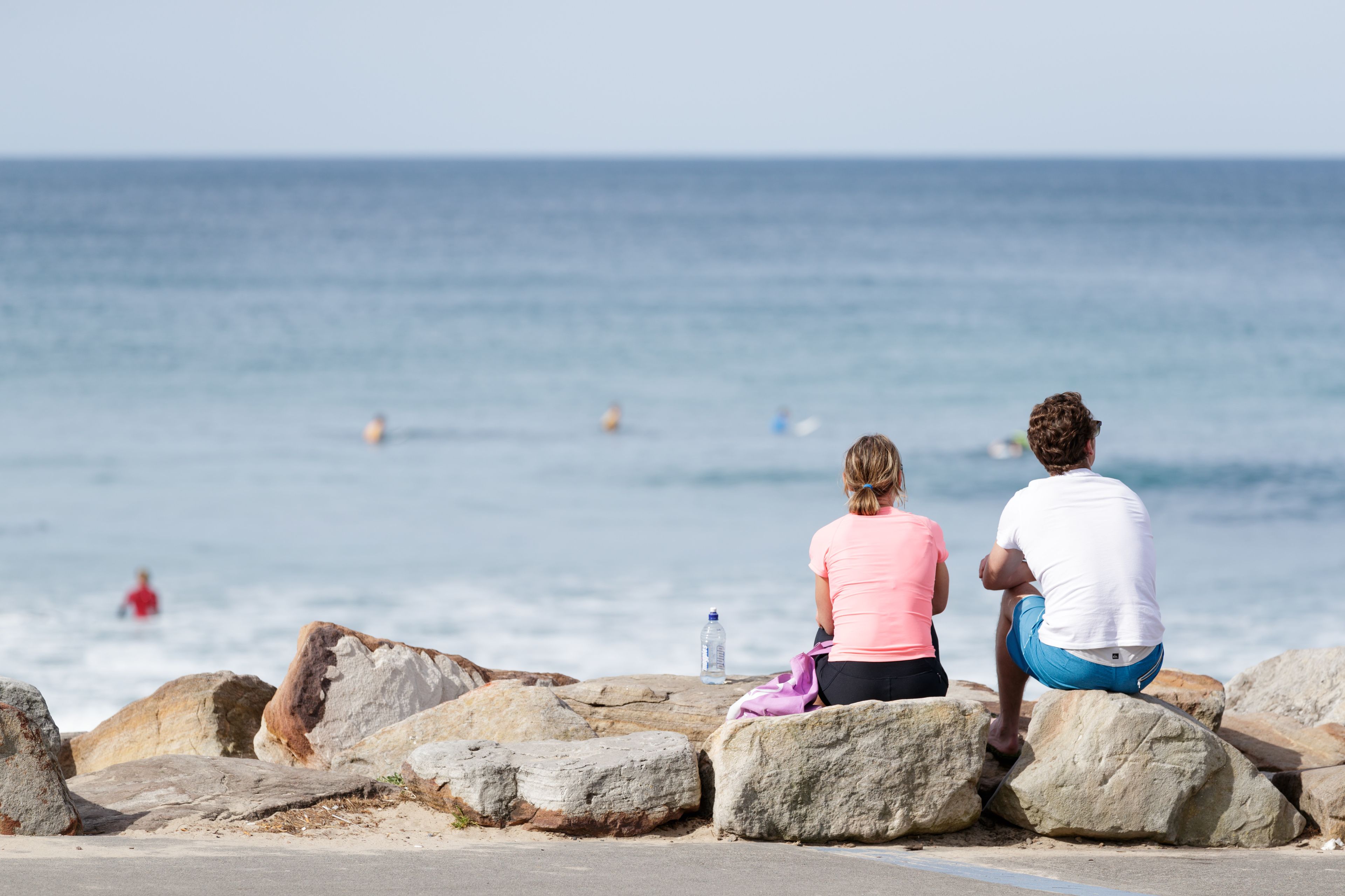 Dos personas sentadas sobre rocas junto al mar, con una botella y una toalla sobre las rocas.