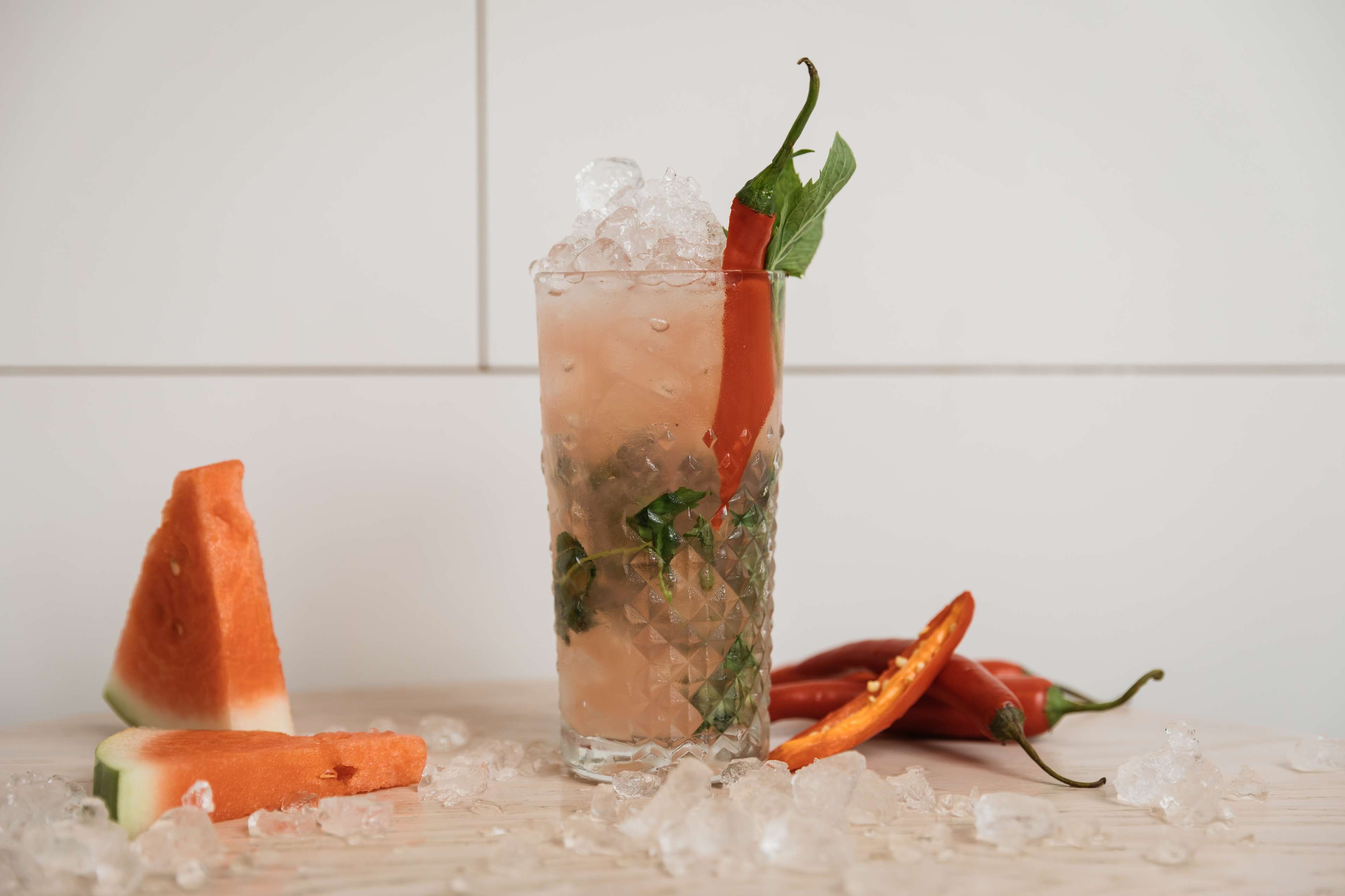 A clear glass with ice cubes, garnished with a red chili pepper and green leaves, placed on a surface with scattered ice cubes and a halved watermelon.