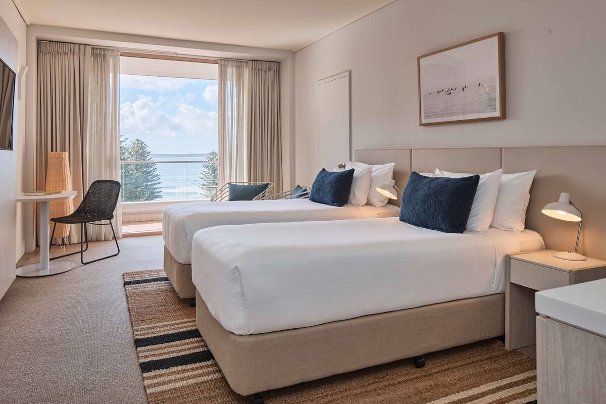 Een hotelkamer met twee bedden, een bureau, een stoel en een groot raam met uitzicht op zee.