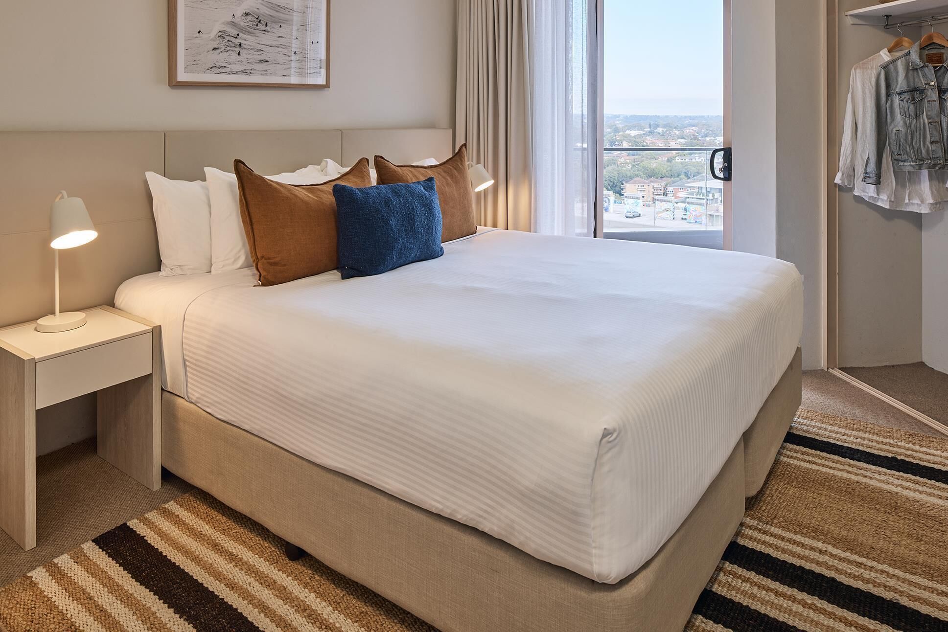 A hotel room with a large bed, a nightstand with a lamp, a view of the city through the window, and a hanging coat rack.
