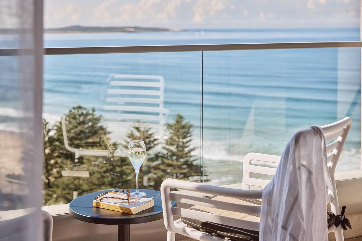Een balkon met uitzicht op zee, een tafel met een boek en een glas, en een witte stoel.