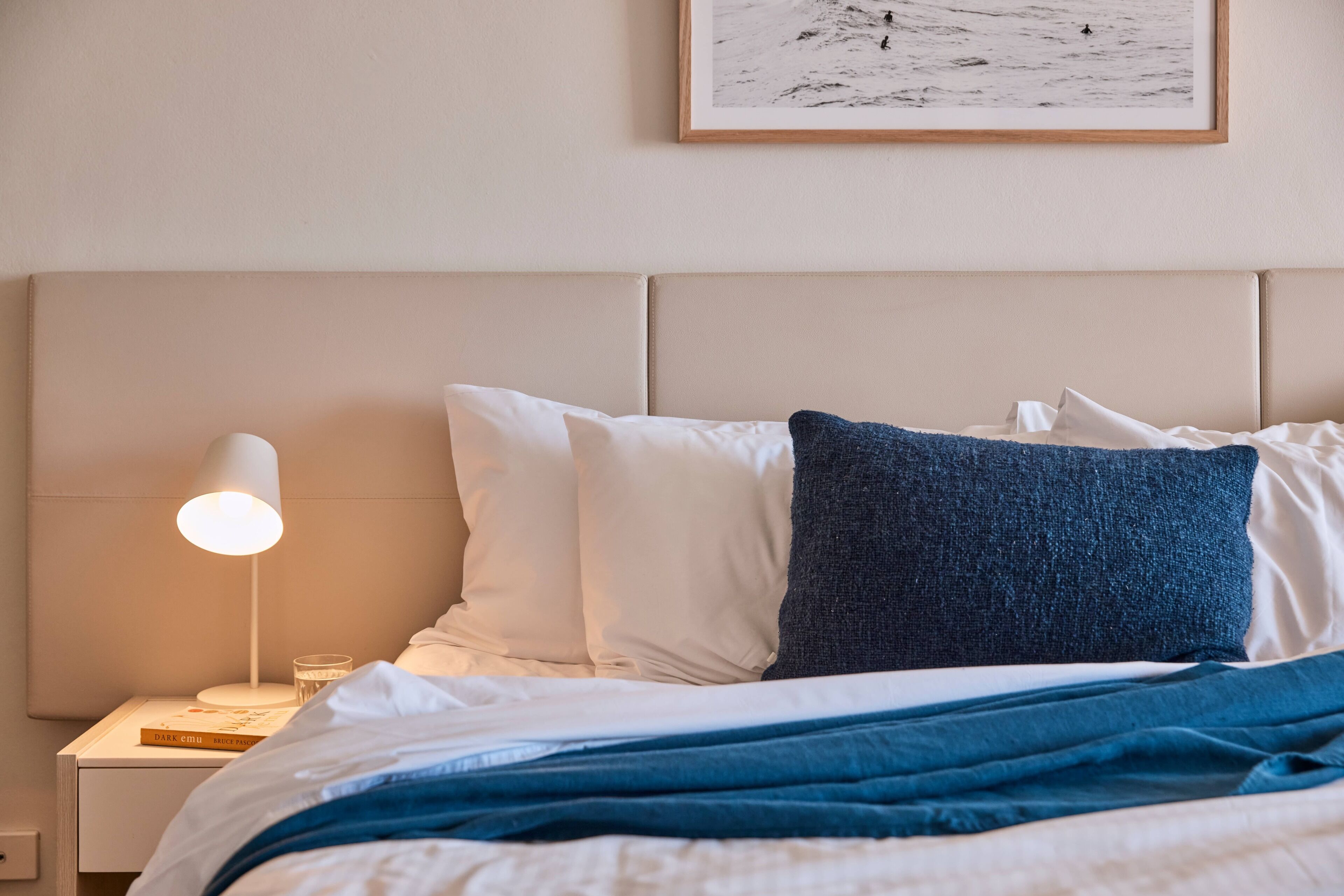 Een net opgemaakt bed met witte en blauwe kussens, een blauwe deken en een bedlampje op een nachtkastje met een boek en een glas.