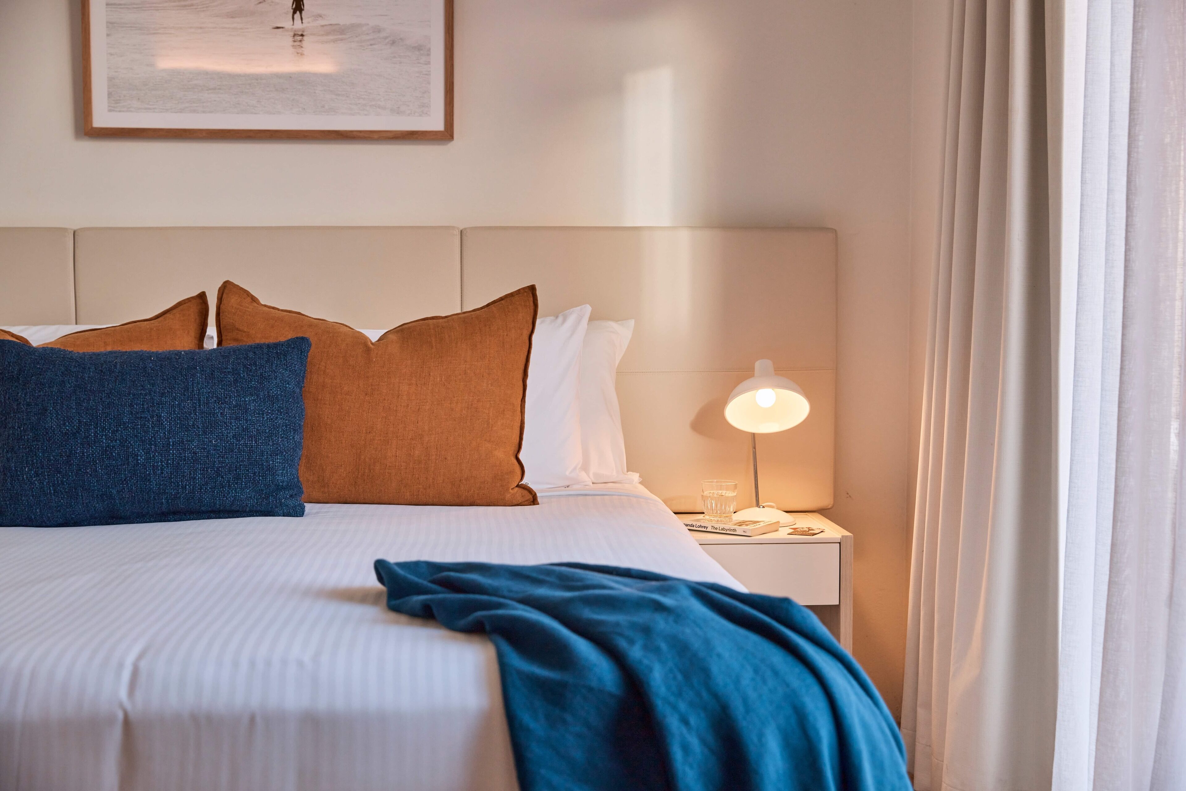 A neatly made bed with a blue blanket, a white pillow, and a brown pillow. There is a nightstand with a lamp and a glass of water.