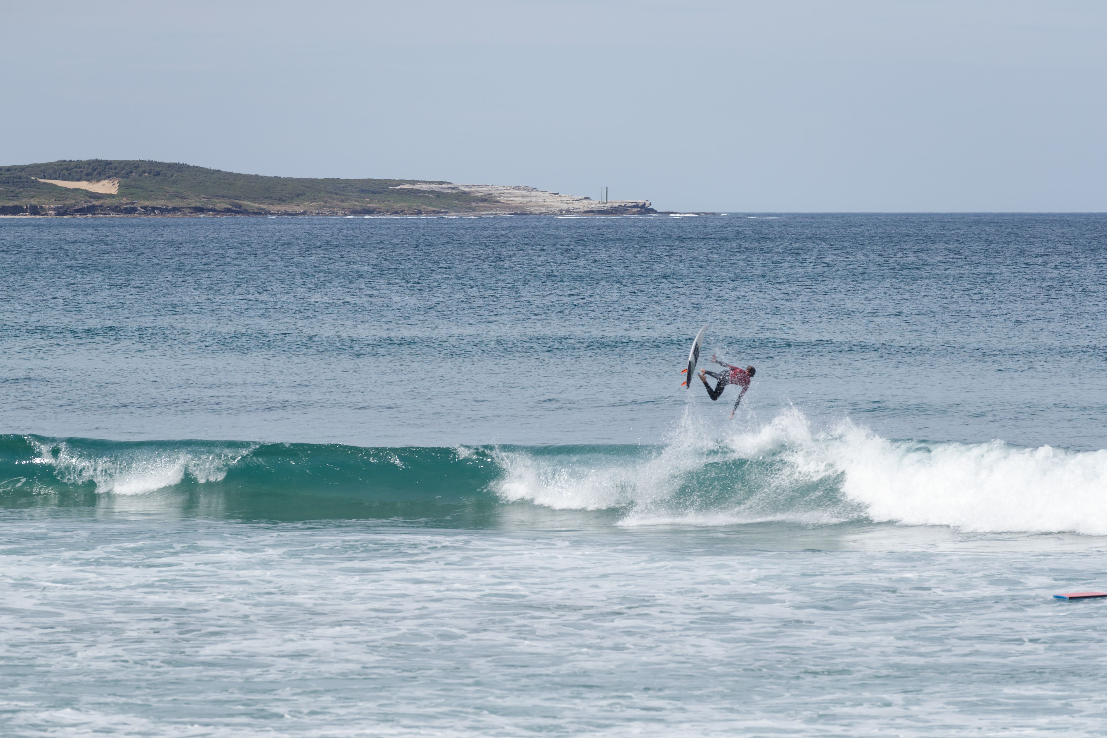 Un surfista cabalgando una ola en el océano, con una costa lejana y cielo despejado.