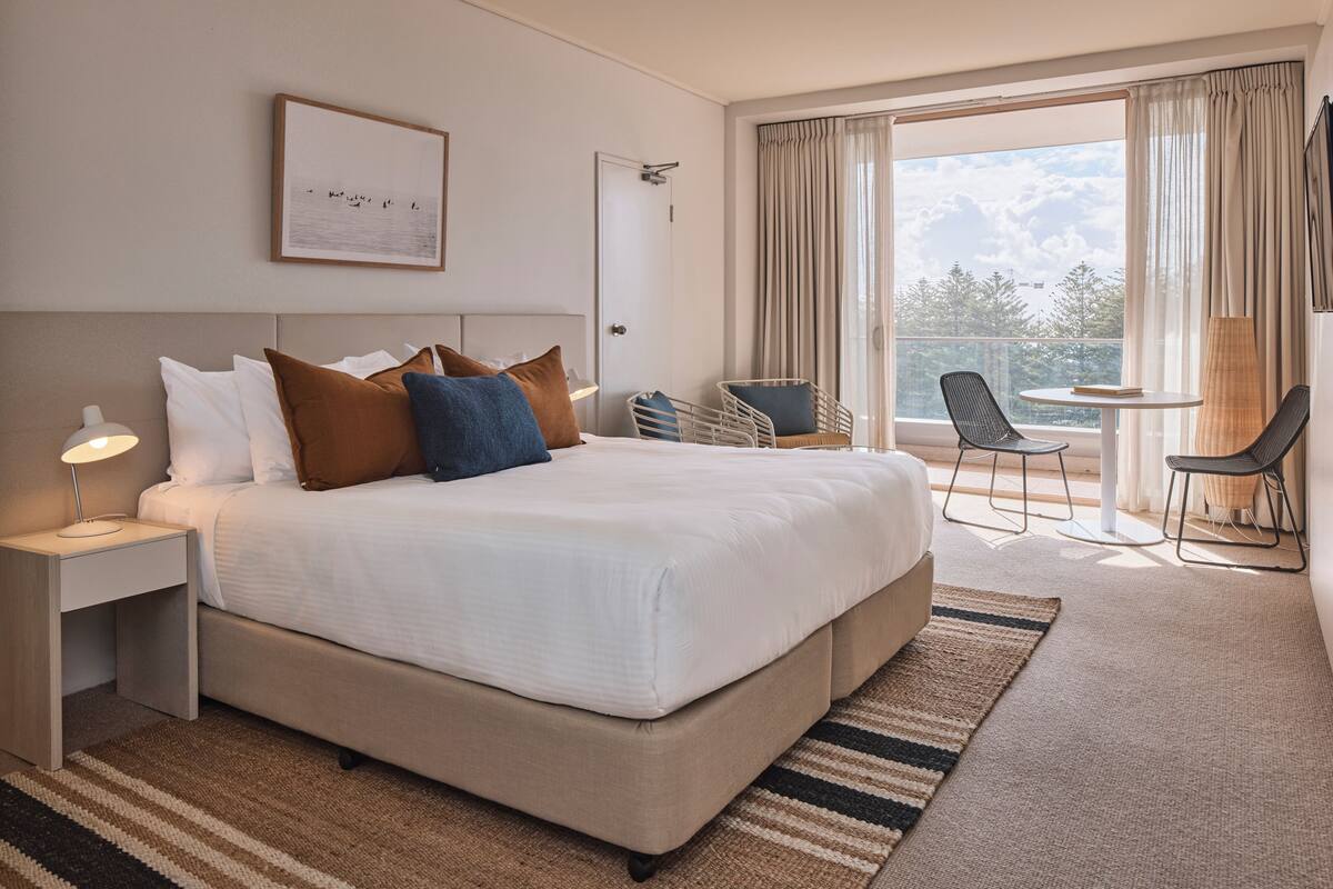 Een moderne hotelkamer met een groot bed, een bureau met lamp, een stoel, een klein tafeltje en uitzicht op het groen.