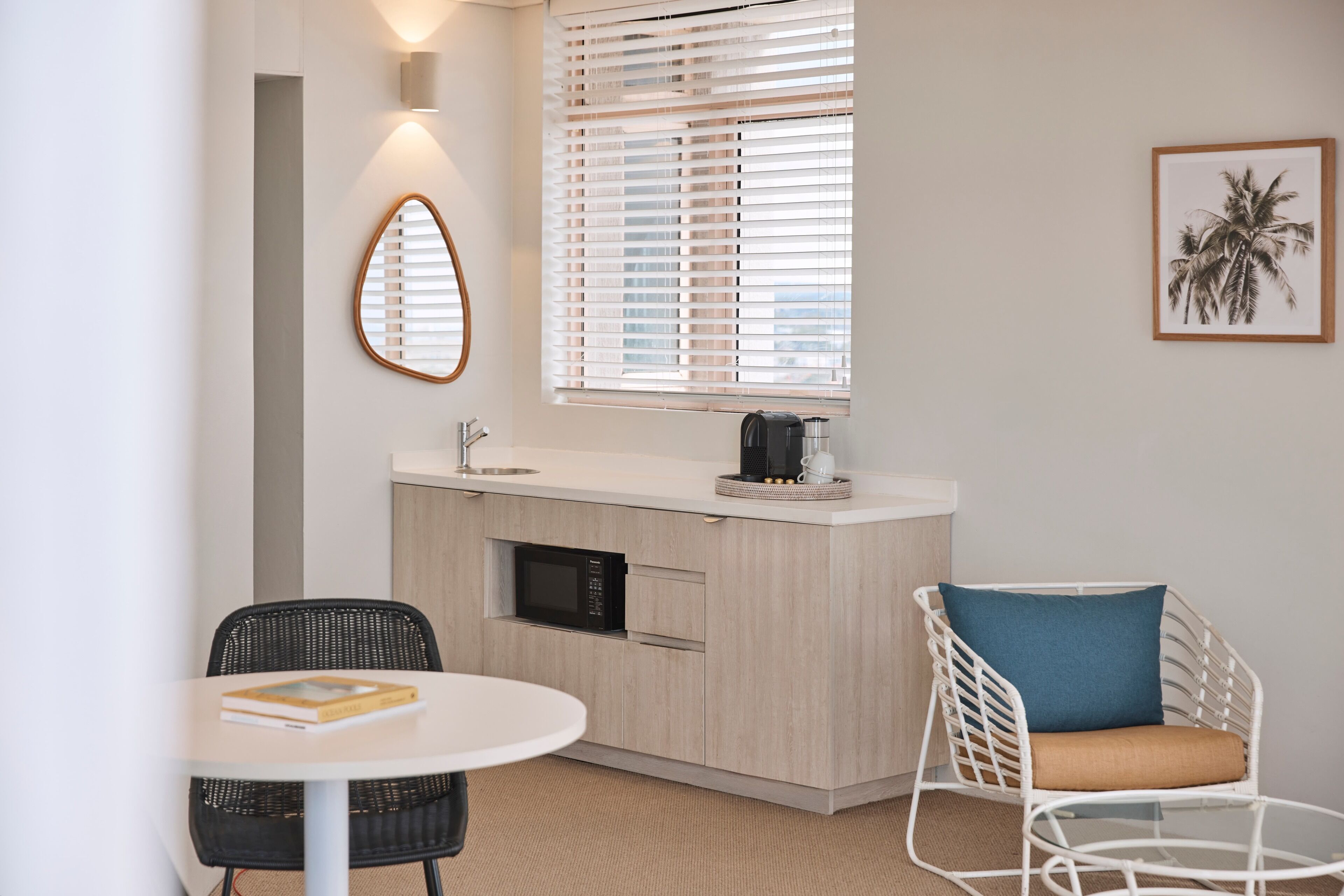 A compact kitchen with a microwave, a coffee maker, and a small sink. There is a round table with a book and a chair with a cushion.