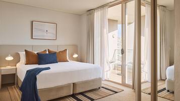 A hotel room with a large bed, a bedside table with a lamp, a framed picture on the wall, and a view through a glass door to an outdoor balcony.