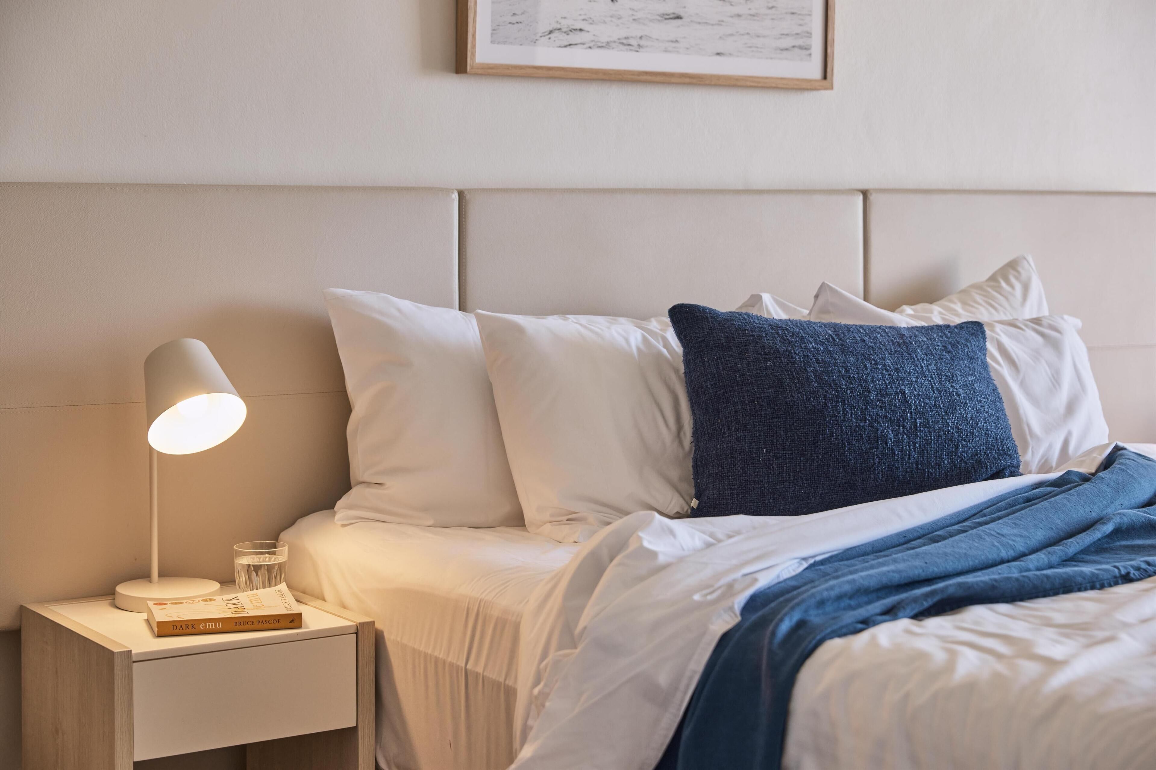 A neatly made bed with white and blue bedding, a bedside lamp, and a small table with a book and glass.