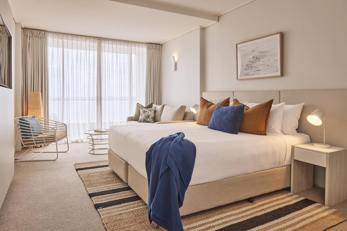 Een moderne hotelkamer met een groot bed, een nachtkastje met een lamp, een stoel en uitzicht op het groen door dunne gordijnen.