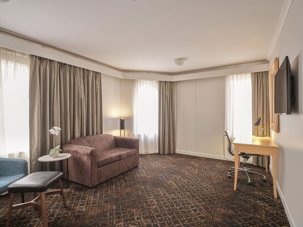 Ein Hotelzimmer mit einer Couch, einem Schreibtisch, einem Sessel, einem kleinen Tisch mit einer Orchidee und einem an der Wand befestigten Fernseher.