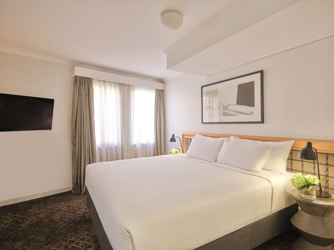 A hotel room with a large bed, a flat-screen TV mounted on the wall, a bedside table with a lamp, and a framed picture above the bed.