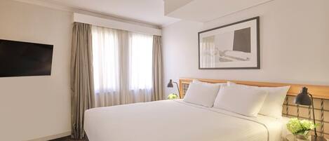 A hotel room with a large bed, a flat-screen TV mounted on the wall, a bedside table with a lamp, and a framed picture above the bed.