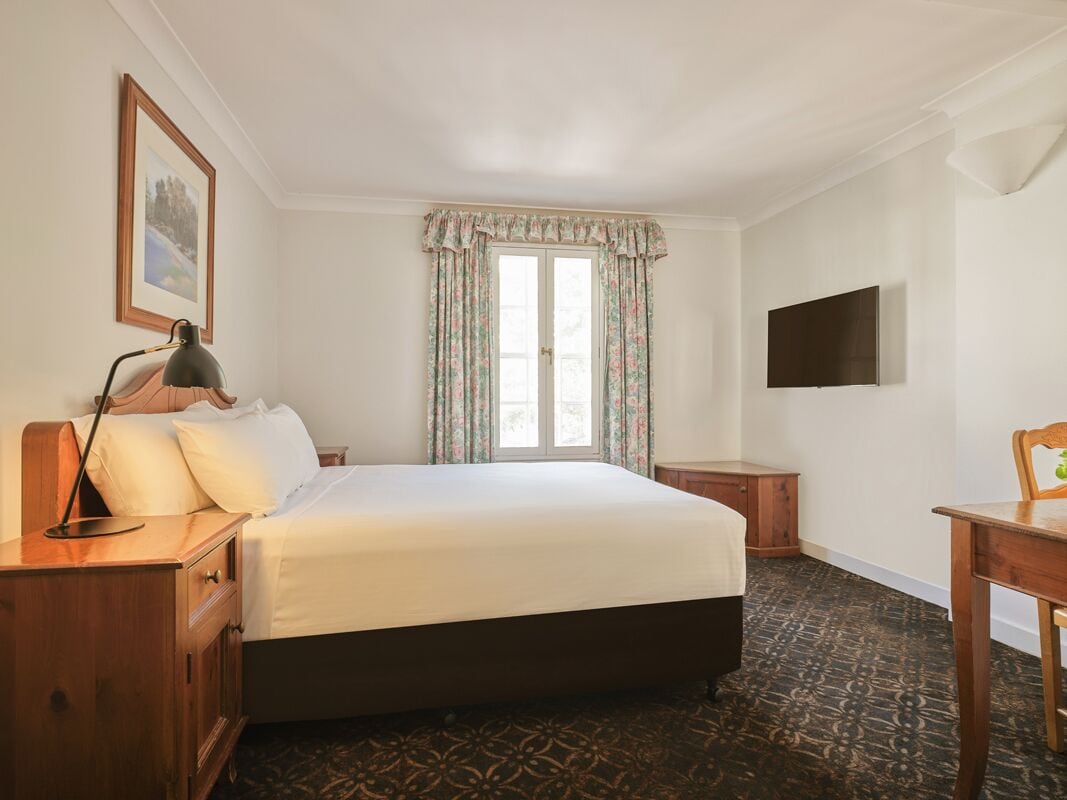Une chambre d’hôtel avec un grand lit, un bureau, une chaise, un téléviseur fixé au mur et une fenêtre avec des rideaux.
