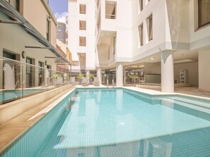 A swimming pool with lounge chairs and a glass railing, surrounded by modern buildings.