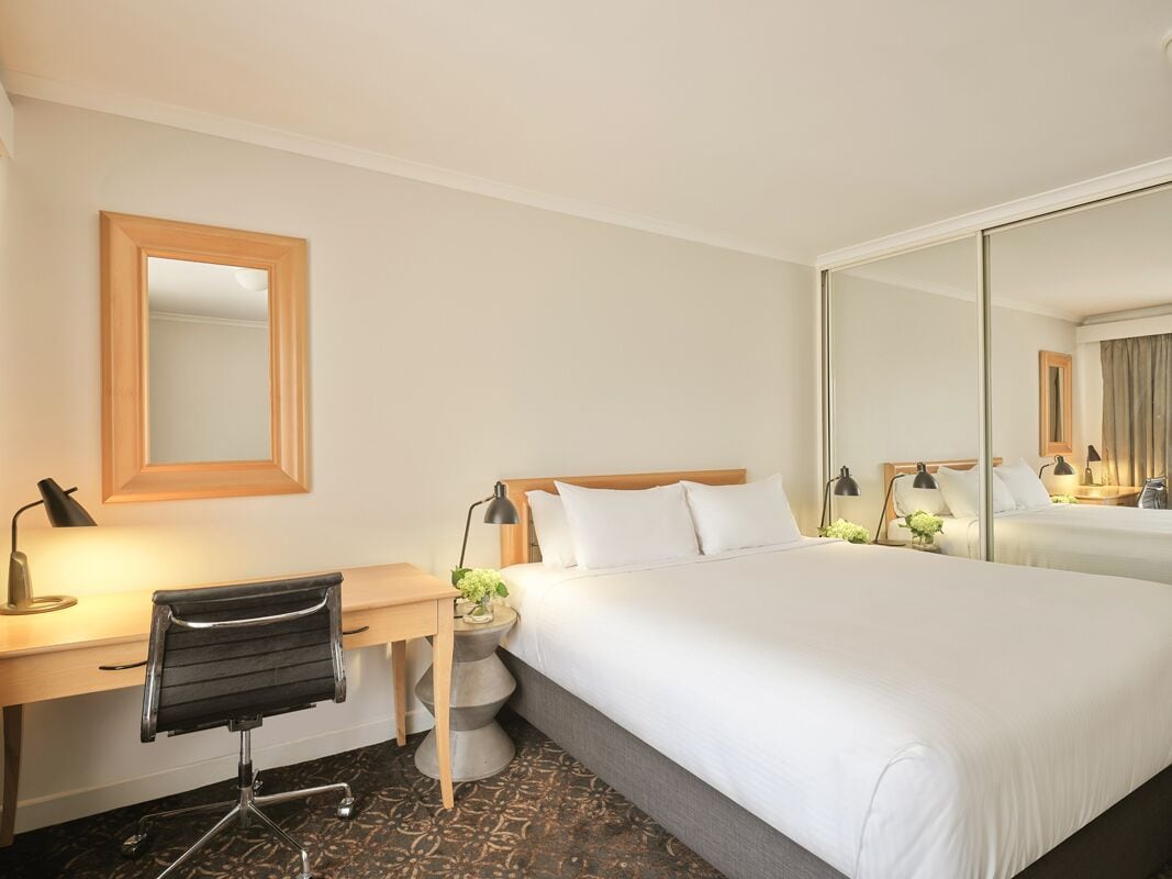 Una habitación de hotel con cama, escritorio, silla, espejo y mesita de noche con lámpara.
