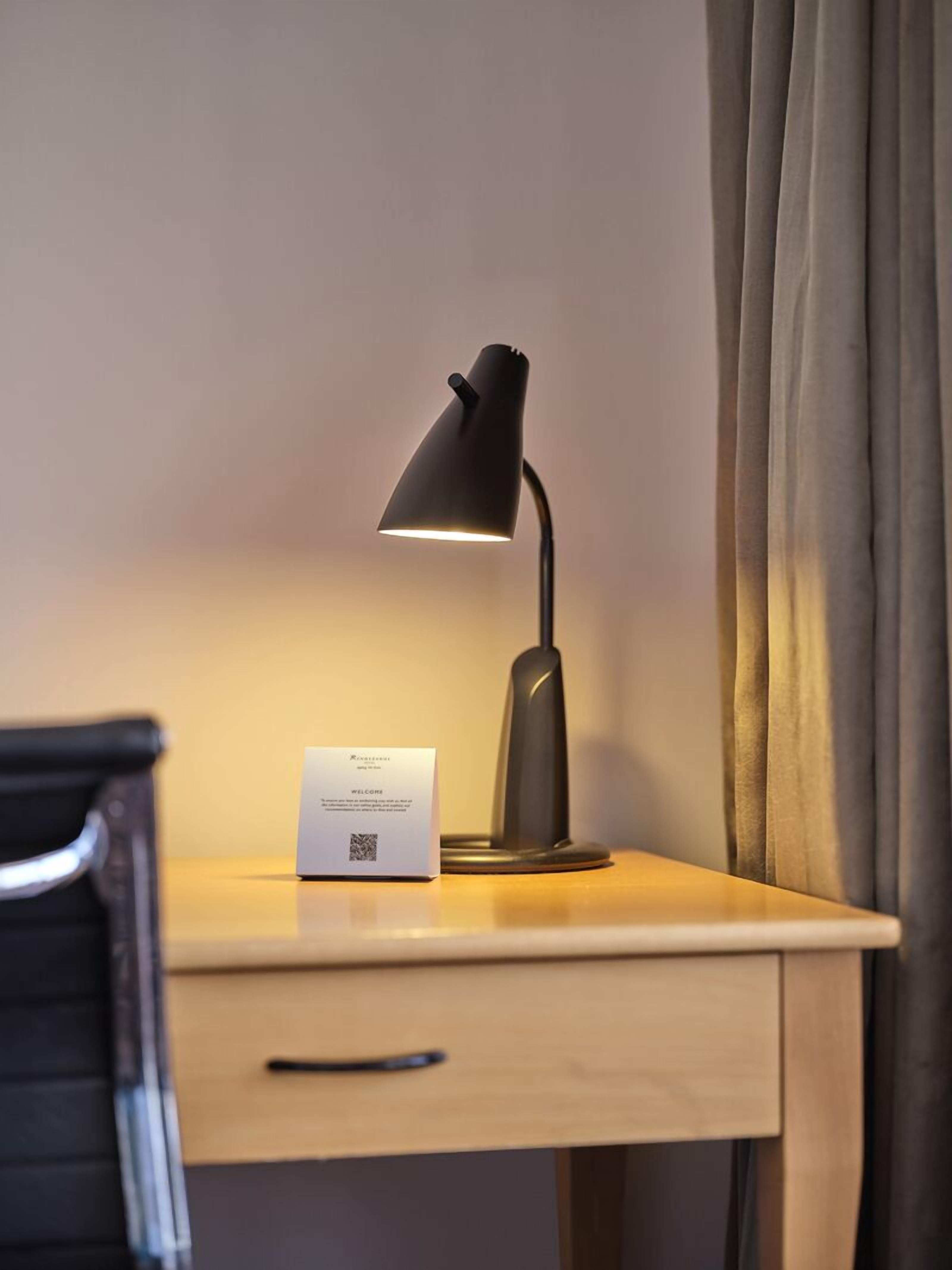 Ett skrivbord med en svart skrivbordslampa, ett vitt välkomstkort och en svart stol.