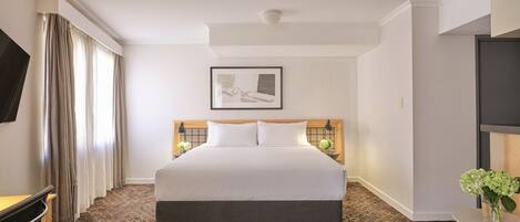 A hotel room with a large bed, a desk, a chair, a TV, and a small table with flowers.