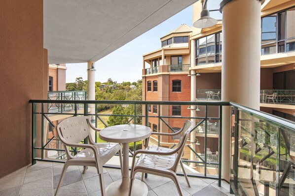 Un balcón con dos sillas blancas y una pequeña mesa redonda, con vistas a un edificio que tiene varios balcones y una vista despejada del entorno.