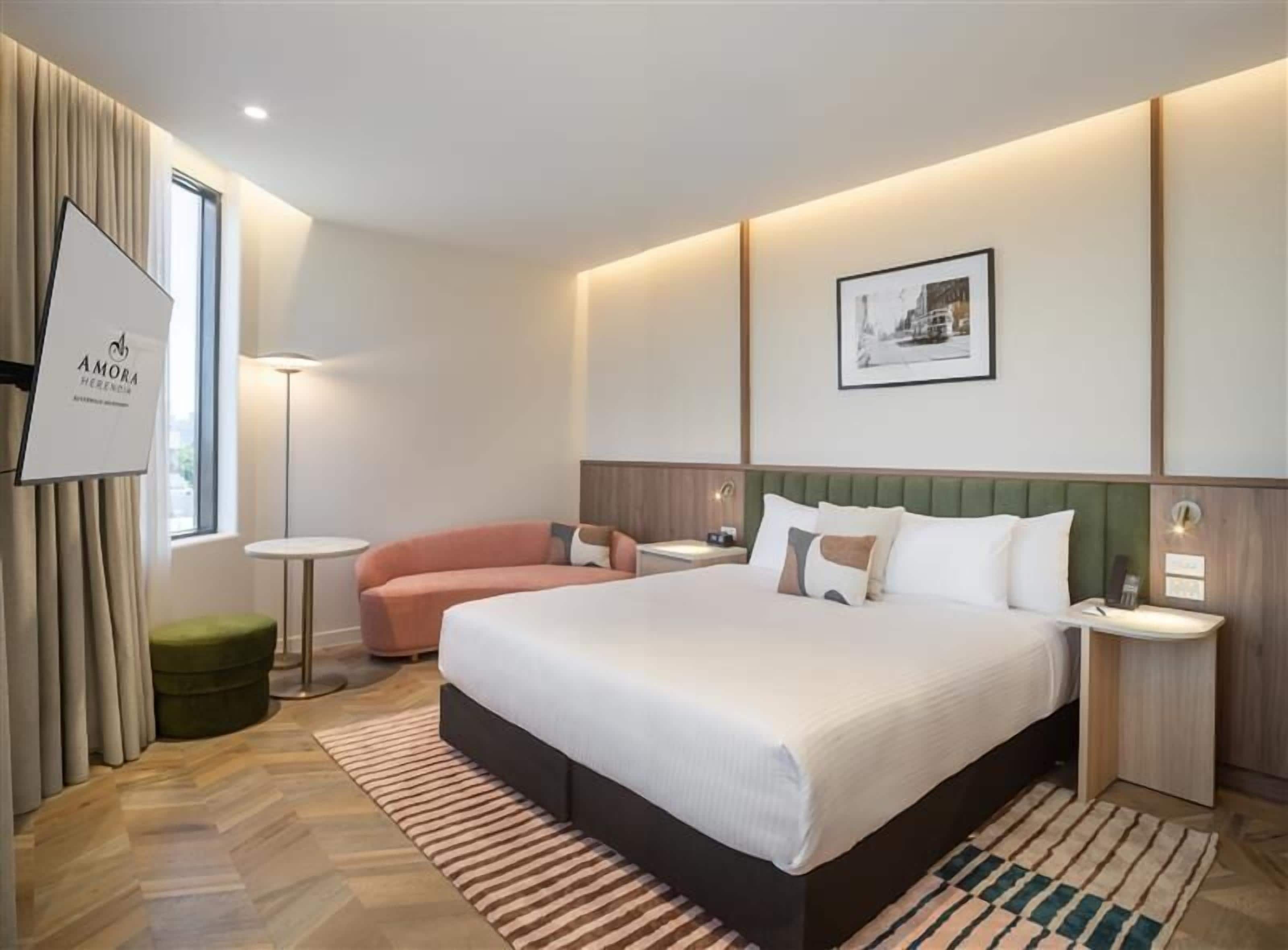 A hotel room with a large bed, a green ottoman, a small round table, a standing lamp, and a framed picture on the wall.