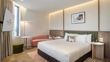 Una habitación de hotel con una cama grande, un reposapiés verde, una mesita redonda pequeña, una lámpara de pie y un cuadro enmarcado en la pared.