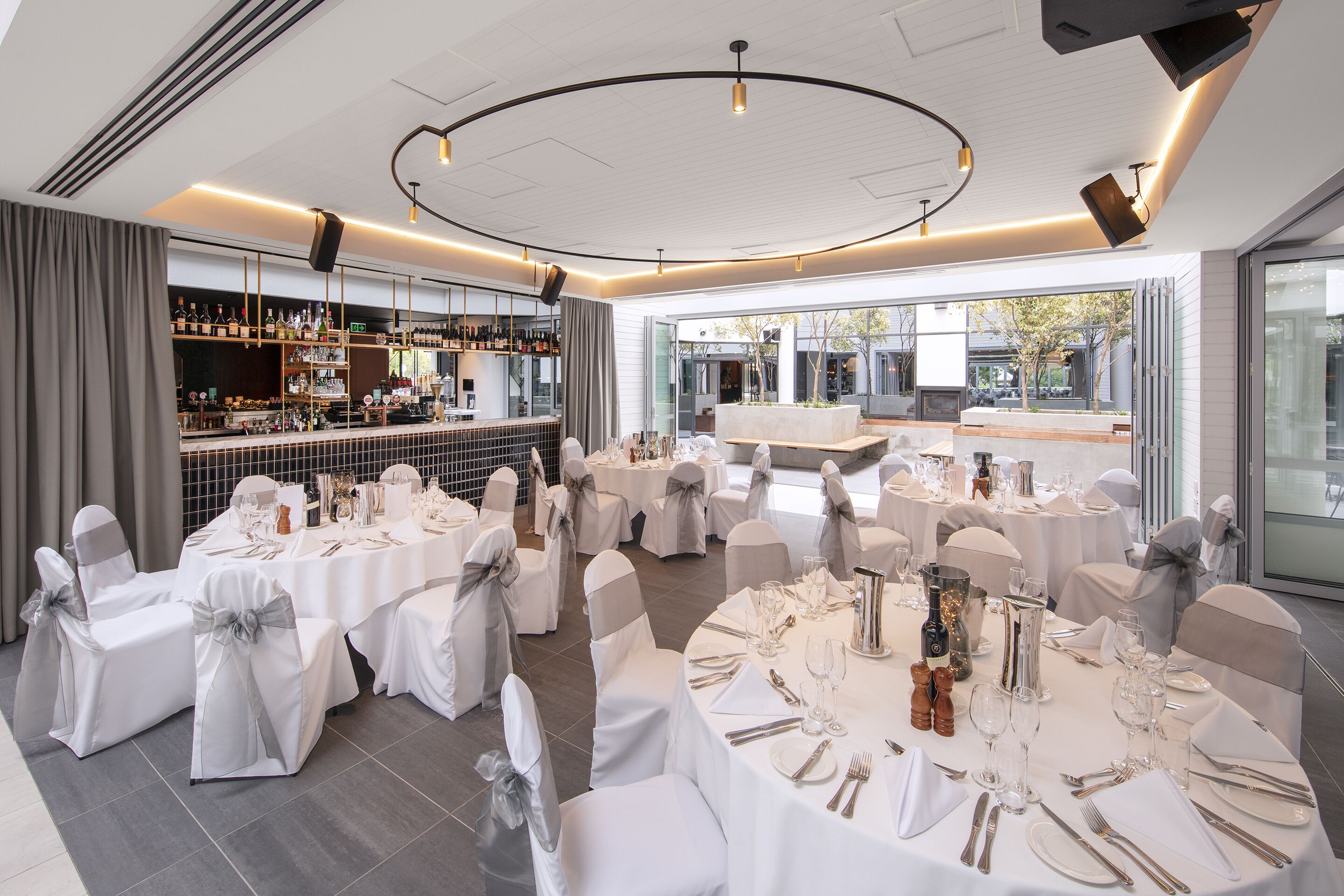 Een feestzaal met ronde eettafels, witte stoelen met grijze linten, een bar met flessen en een uitzicht op het buitengebied door grote ramen.