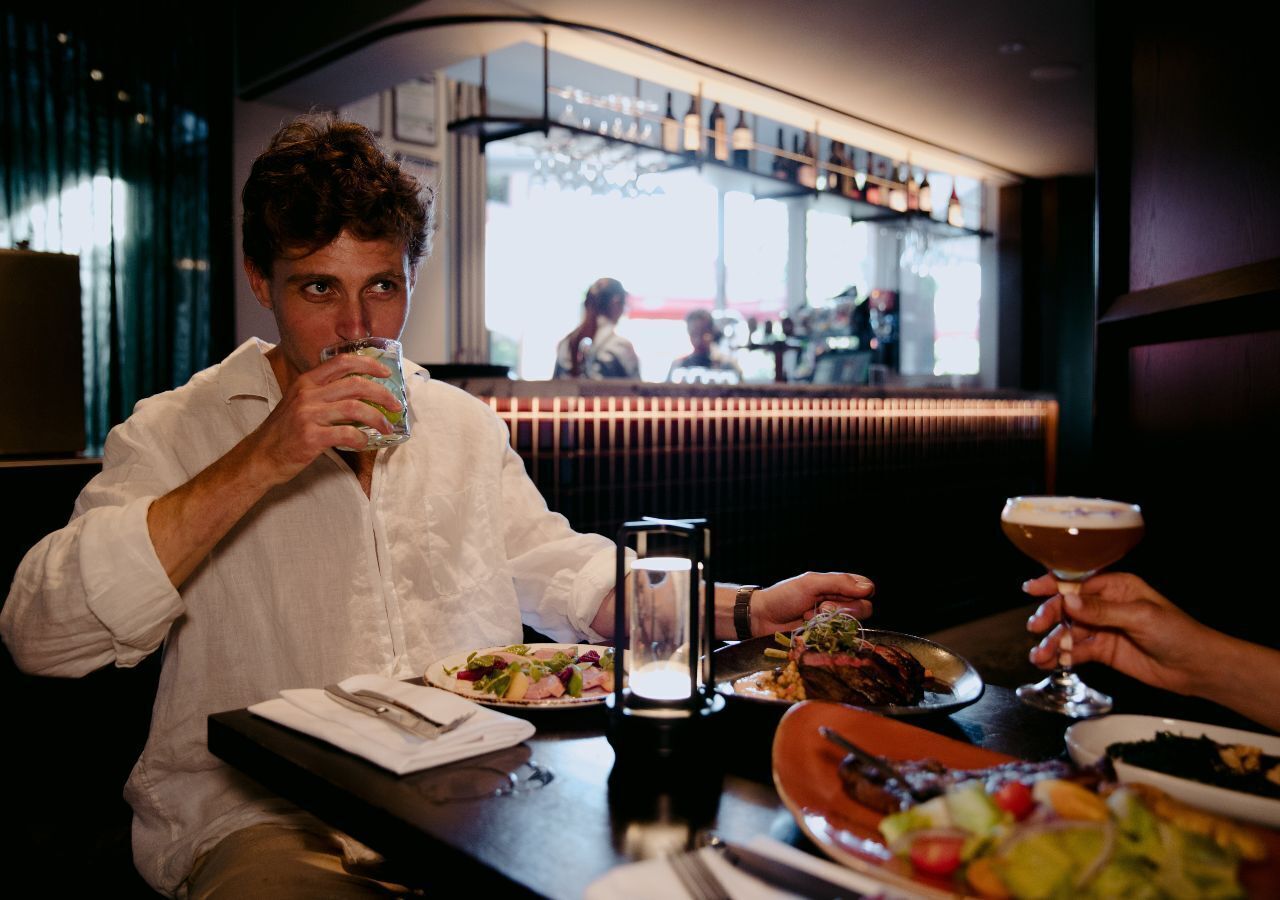 A person seated at a table with a drink, a candle, and various dishes in a dimly lit restaurant with a bar in the background.