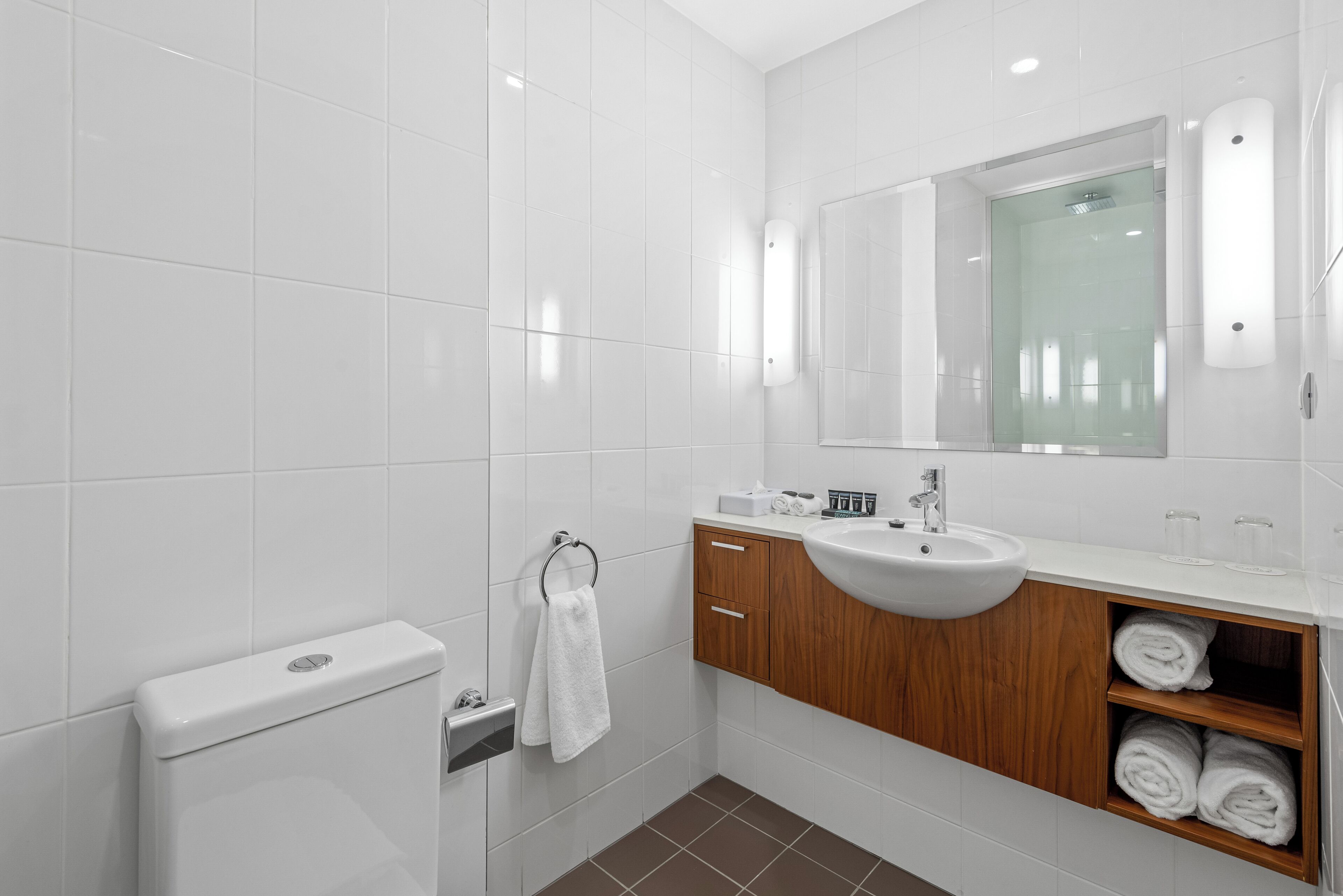 A modern bathroom with white tiles, a wooden vanity, a sink, a toilet, and a towel rack.