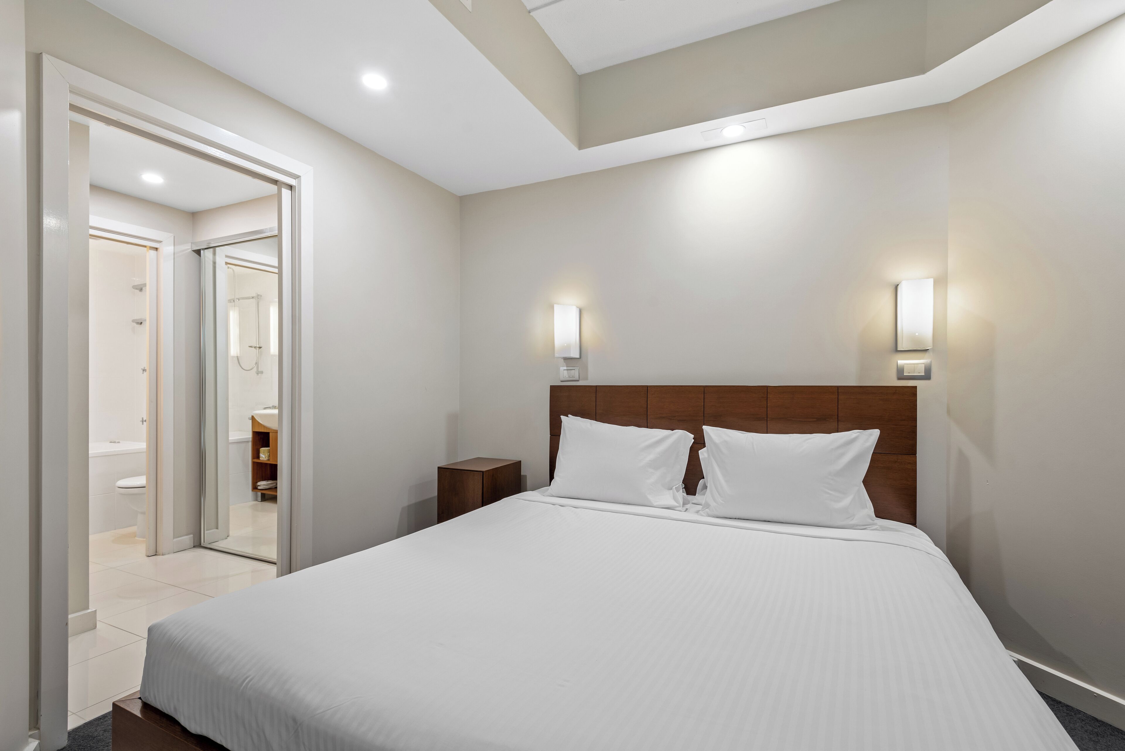 A modern hotel room with a large bed, a wooden headboard, and two wall-mounted lights.