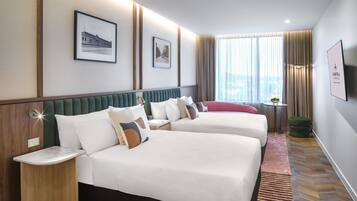 A hotel room with two beds, a green upholstered headboard, a small table with a lamp, a TV mounted on the wall, and a window with curtains.