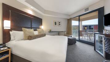 A hotel room with a large bed, a desk with a chair, a balcony with a table and chairs, and a TV mounted on the wall.