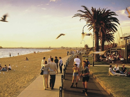 A beachside walkway with people walking and sitting, palm trees, and a view of the ocean.. Mercure Welcome Melbourne