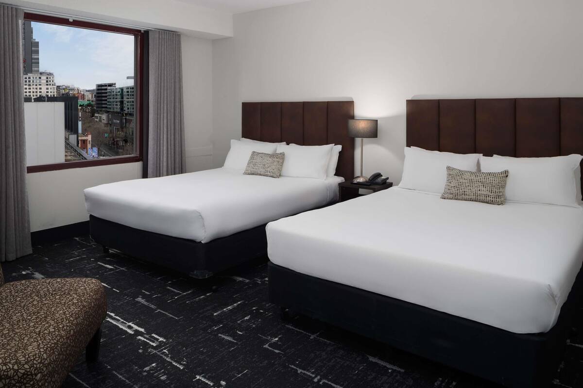 A hotel room with two beds, a nightstand with a lamp, a chair, and a view of the city through the window.
