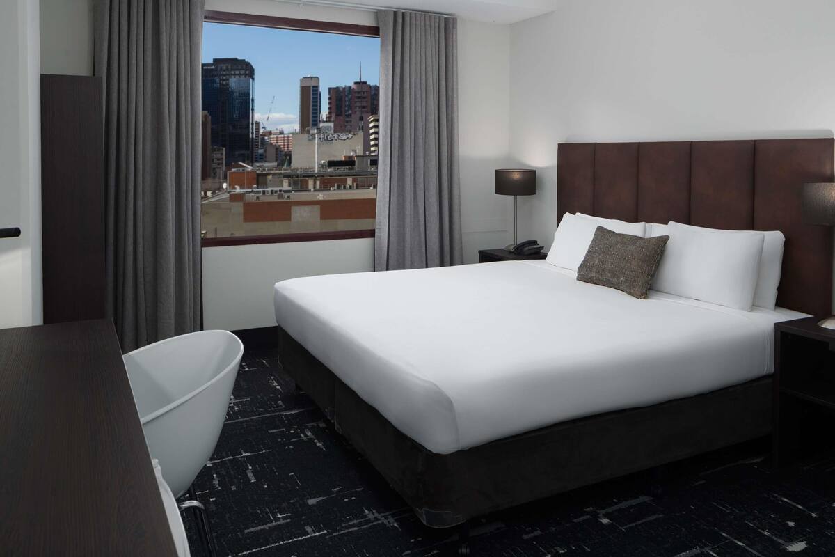 A hotel room with a large bed, a desk, a chair, a nightstand with a lamp, and a view of the cityscape through the window.