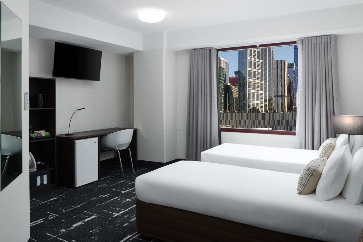 A hotel room with a bed, a desk, a chair, a TV, and a window with a city view.