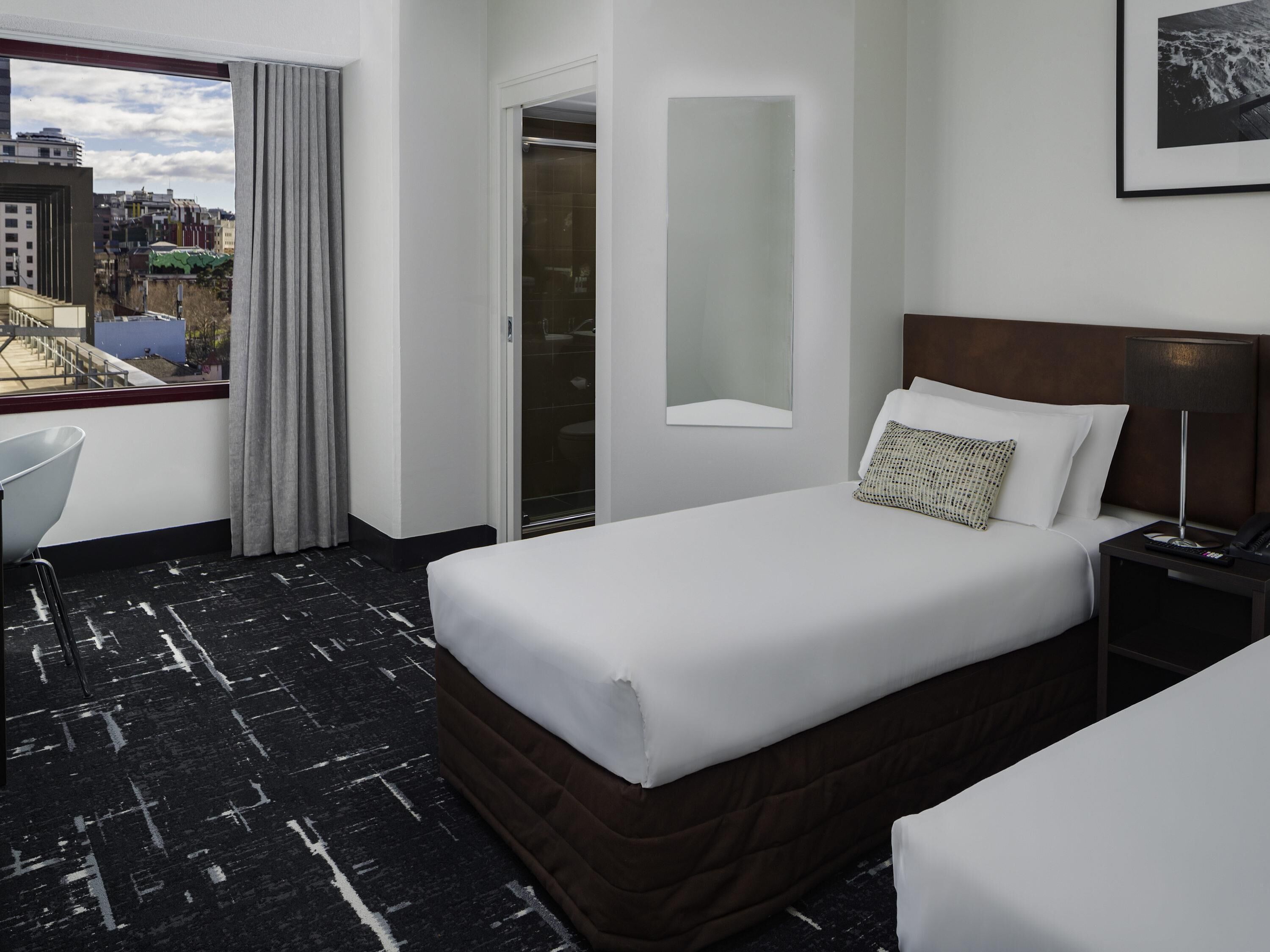 A hotel room with a bed, a desk, a chair, a window with curtains, a mirror, and a city view.
