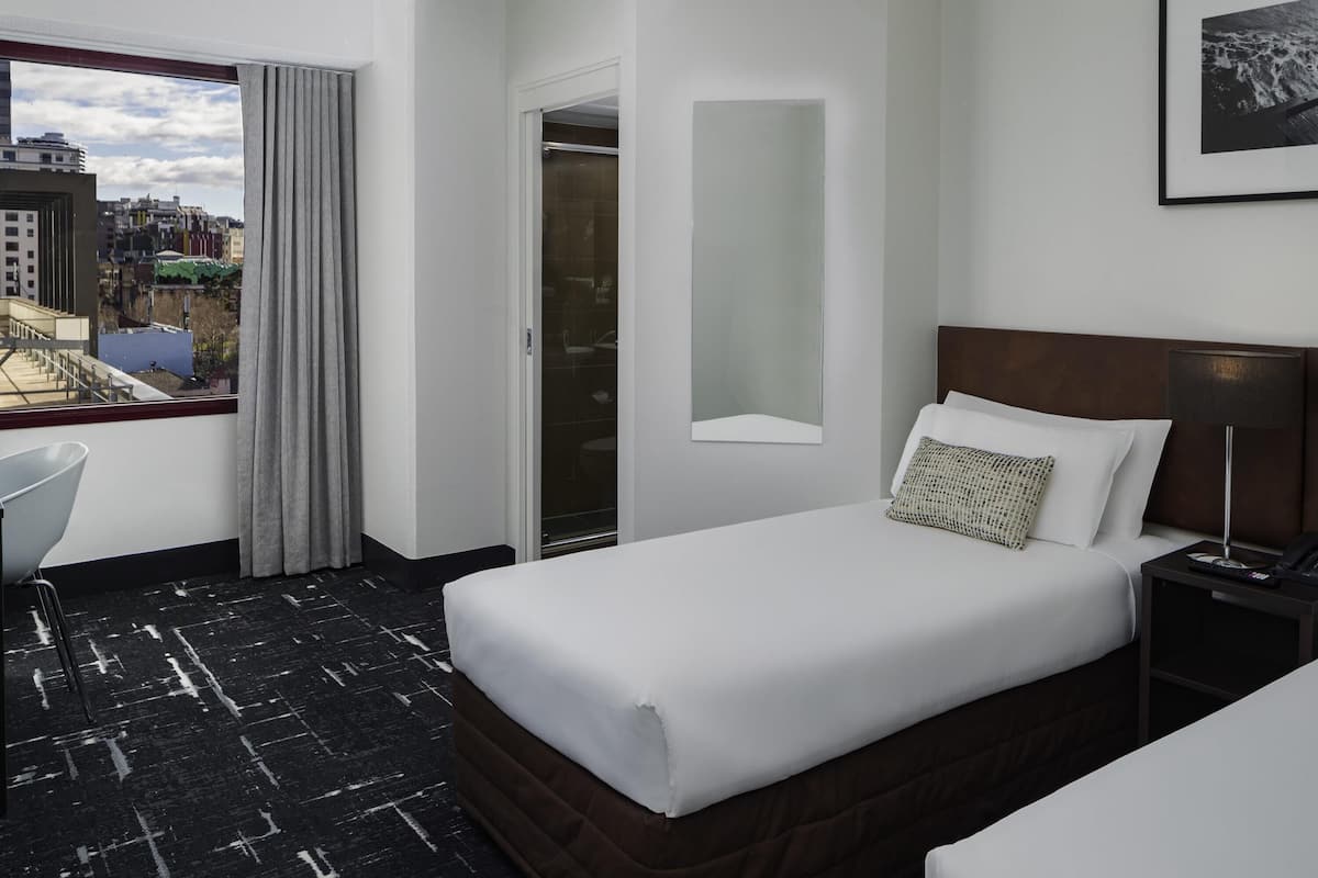 A hotel room with a bed, a desk, a chair, a window with curtains, a mirror, and a city view.