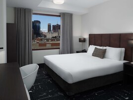 A hotel room with a large bed, a desk, a chair, a nightstand with a lamp, and a view of the cityscape through the window.