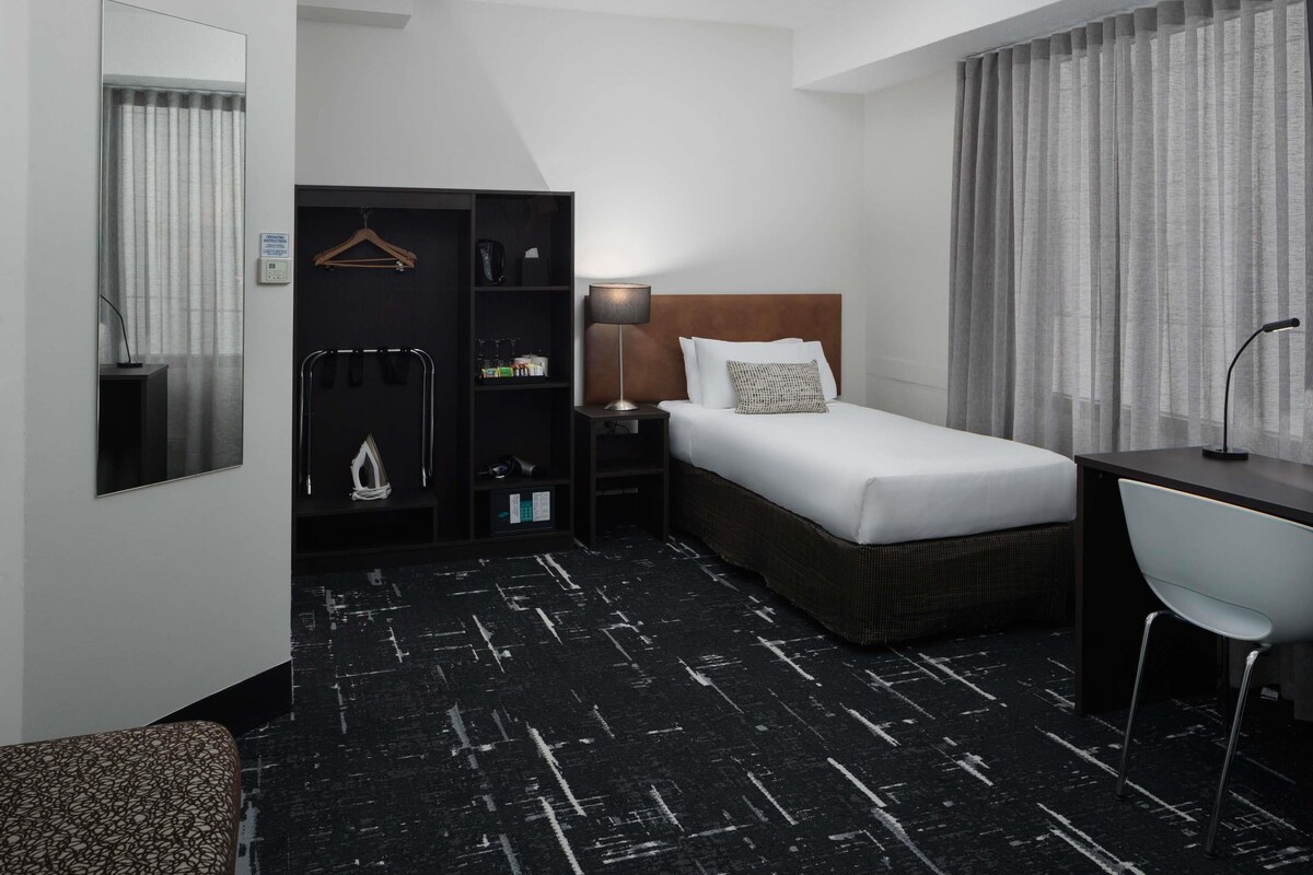 Habitació d'hotel amb un llit, una taula de nit, una cadira, un armari i un mirall.