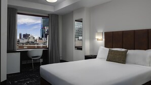Habitación de hotel con una cama grande, un escritorio y una silla. Se ve un paisaje urbano a través de la ventana.