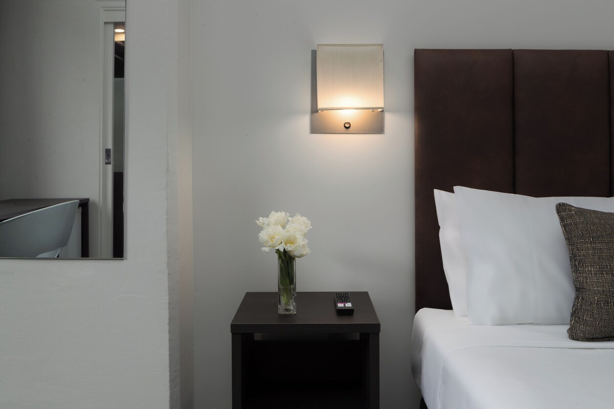 Habitación de hotel con una cama, una mesita de noche con un florero, un control remoto y una lámpara fijada en la pared.