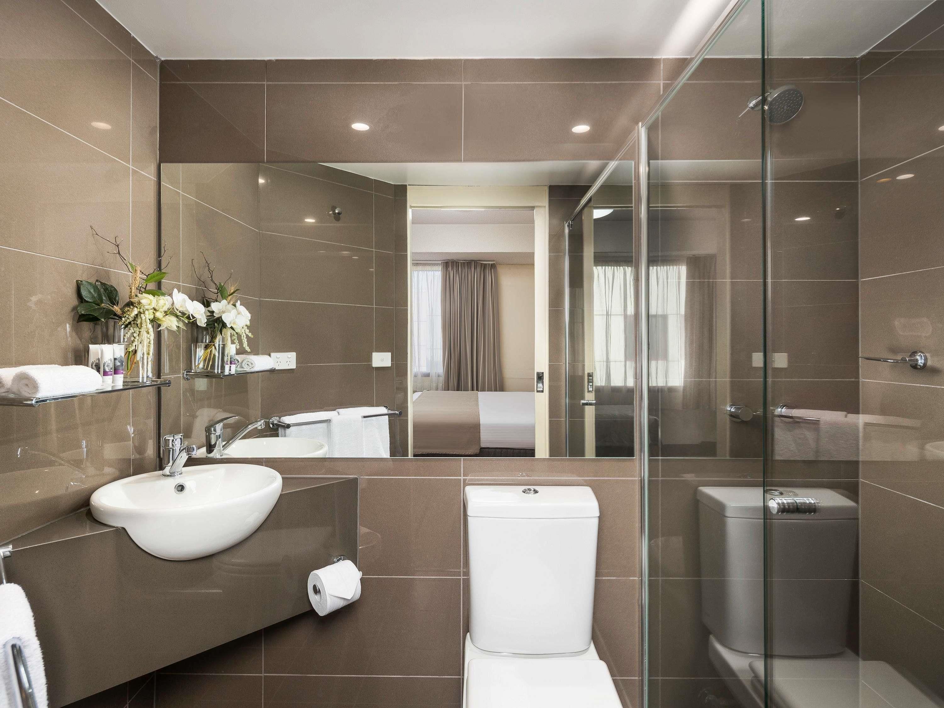 Un baño moderno con cabina de ducha de vidrio, un espejo grande y una habitación visible al fondo.