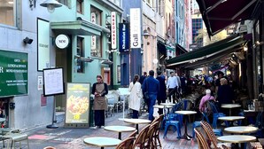 Un café al aire libre con mesas y sillas, rodeado de edificios con diversos letreros y escaparates.
