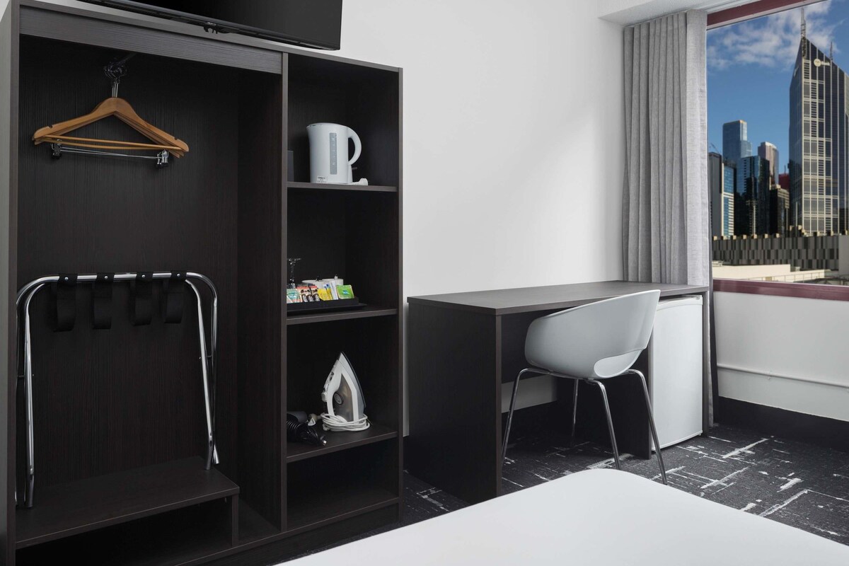 Habitació d'hotel moderna amb un armari de fusta fosca, un escriptori, una cadira blanca i vista a la ciutat a través d'una finestra gran.