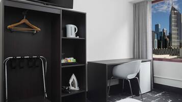 Habitación de hotel moderna con un armario de madera oscura, un escritorio, una silla blanca y vistas a la ciudad a través de un ventanal grande.