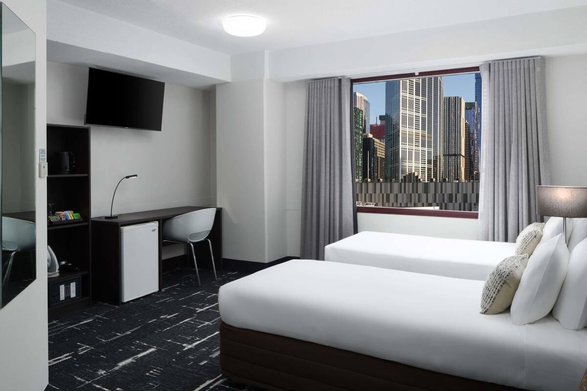 A hotel room with a bed, a desk, a chair, a TV, and a window with a city view.