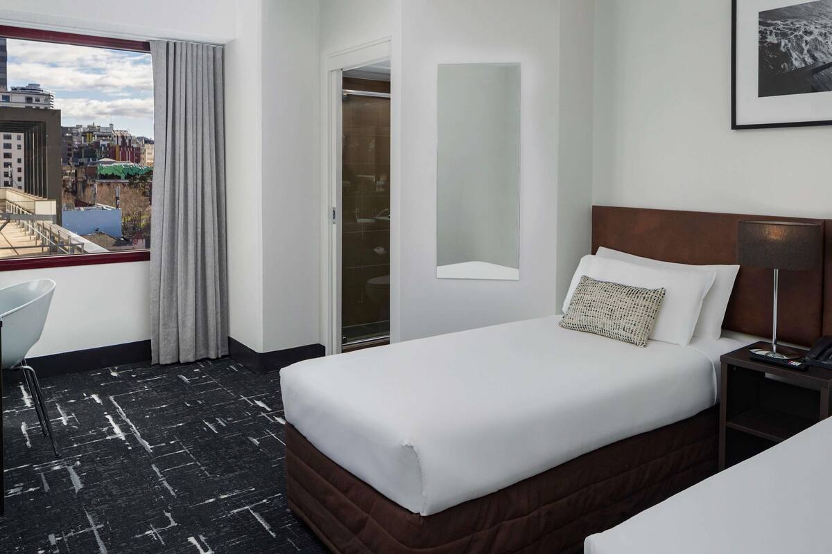 A hotel room with a bed, a desk, a chair, a window with curtains, a bathroom, and a city view.