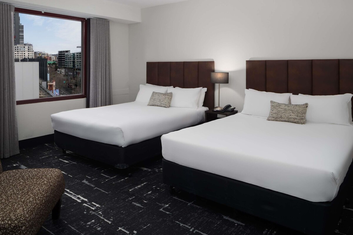 A hotel room with two beds, a nightstand with a lamp, a chair, and a view of the city through the window.